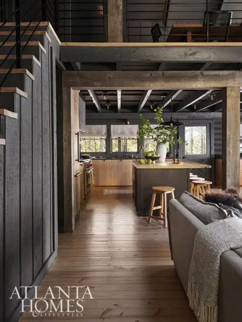 Rustic kitchen and living area with dark wood beams, wooden floors, and a gray sofa.