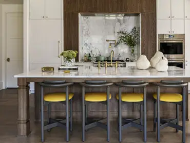 Kitchen island with marble countertop, yellow bar stools, and wood cabinetry. Marble backsplash