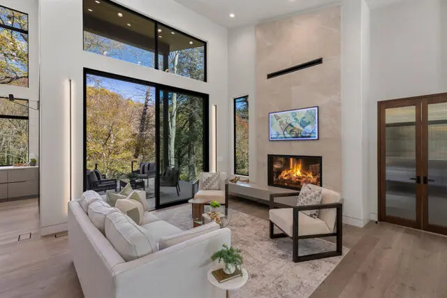 Living room with white sofas, chairs, fireplace, and large windows overlooking autumn trees.