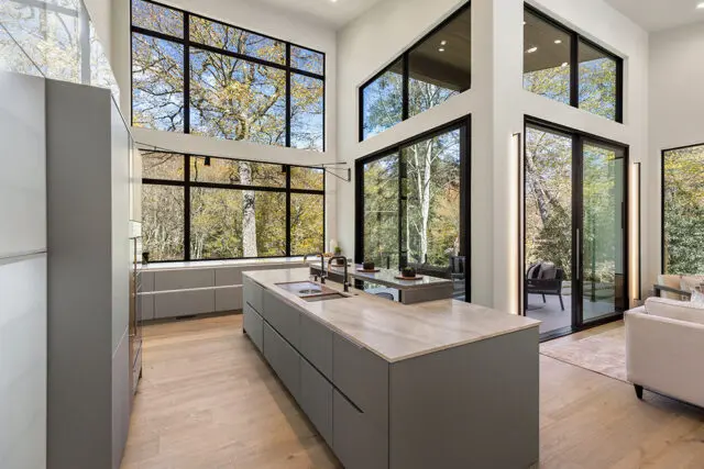 Modern gray kitchen with marble island, large windows overlooking trees, and light wood floors.