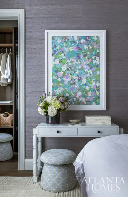 Bedroom with grasscloth wallpaper, floral art, white desk, and patterned ottomans.