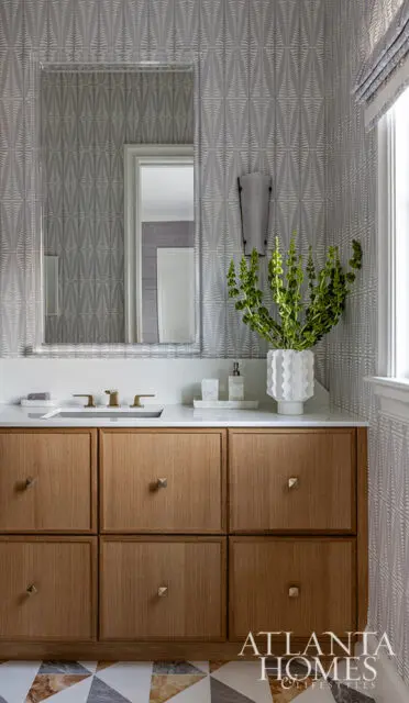 Bathroom vanity with oak cabinets, white countertop, gold faucet, and patterned wallpaper.