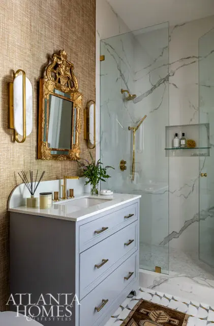 Gray vanity with brass hardware, marble shower, and gold ornate mirror.