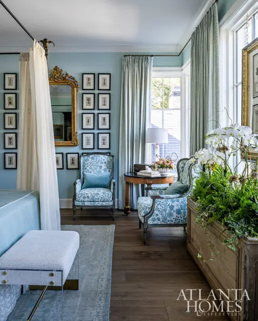 Light blue bedroom with canopy bed, floral armchairs, gold mirror, and botanical prints
