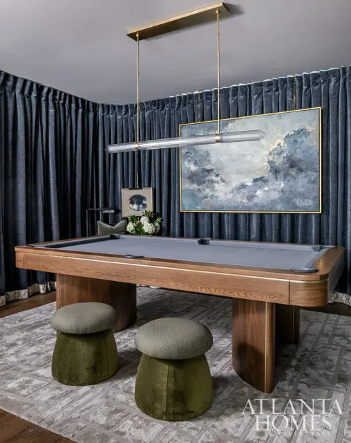 Game room with gray pool table, two olive green stools, and abstract art above dark blue curtains.
