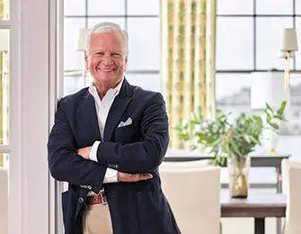 Man in navy blazer and tan pants smiles with arms crossed in a well-lit room.