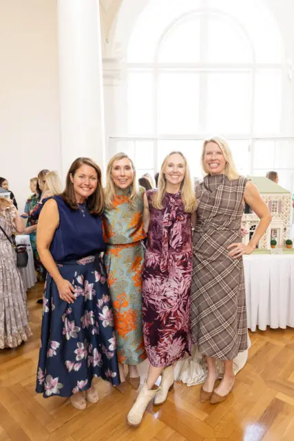 Four women posing for a photo indoors, with a dollhouse in the background.