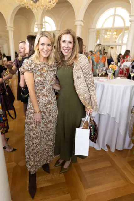 Two women smiling at a party. One wears a floral dress, the other a green dress and textured jacket.