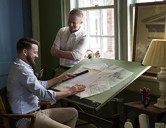 Two men reviewing architectural drawings at a drafting table in a room with green walls and a large