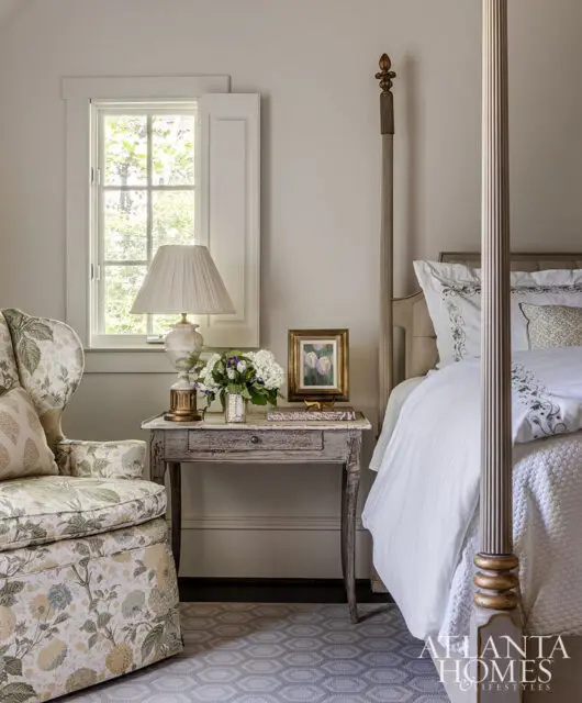 Bedroom with floral armchair, antique side table, and four-poster bed.