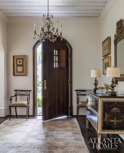 Entryway with arched wooden door, ornate chandelier, antique console table, and two chairs.