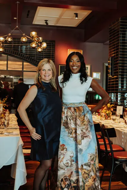 Two women posing in a restaurant setting. One wears a navy dress, the other a white top and ornate