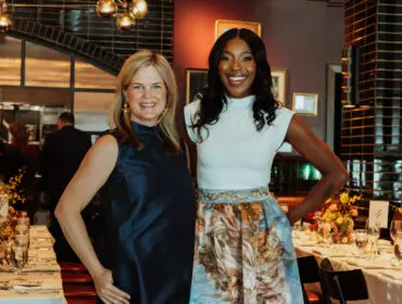 Two women smiling at a formal dinner setting. One wears a navy dress, the other a white top and