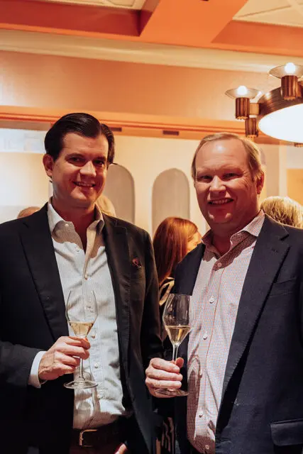 Two men in suits holding champagne flutes, smiling.