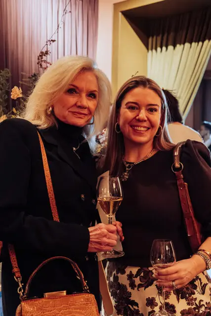 Two women smiling, holding wine glasses. One has blonde hair, the other has brown hair.