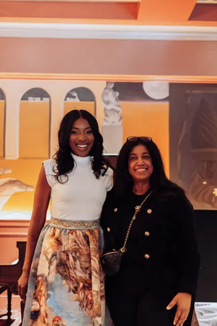 Two women smiling, one in a white top and patterned skirt, the other in a black jacket.
