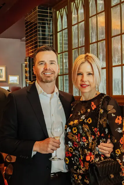 Man in suit holding wine glass, woman in floral dress with purse.
