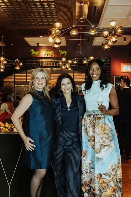Three women smiling, two holding wine glasses, in a room with ornate lighting and decor.