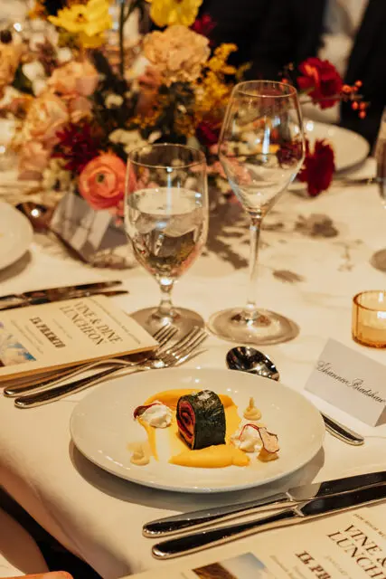White plate with rolled food, yellow sauce, and white dollops. Place setting with wine glasses and