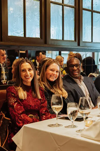 Three people seated at a table with wine glasses. Woman in red lace dress, woman in black sweater