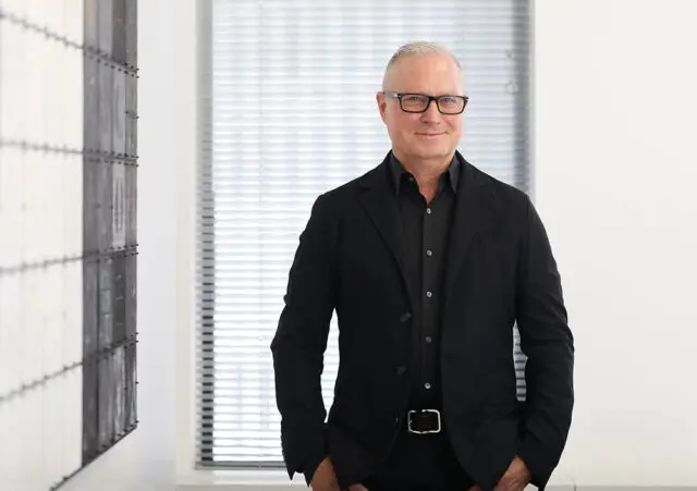 Man in black suit and glasses smiling in front of blinds