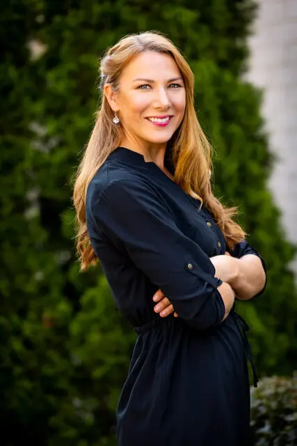 Woman with long blonde hair wearing a black dress with arms crossed, smiling.