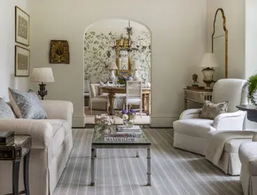 Living room with neutral sofas and chairs, arched doorway to dining room with floral wallpaper.