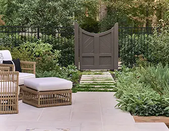Wicker chairs and ottoman on stone patio with garden gate and lush greenery