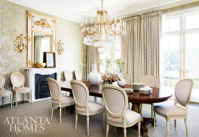 Elegant dining room with a dark wood oval table, upholstered chairs, ornate fireplace, crystal chandelier, and floral wallpaper.