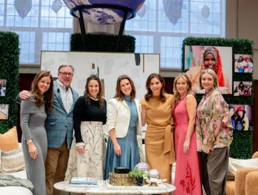 Seven people posing for a photo in front of a backdrop featuring photos and greenery. The group includes four women and three men, all dressed in stylish attire.
