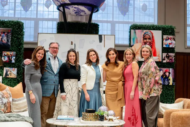 Seven people posing for a photo in front of a backdrop featuring photos and greenery. The group includes four women and three men, all dressed in stylish attire.