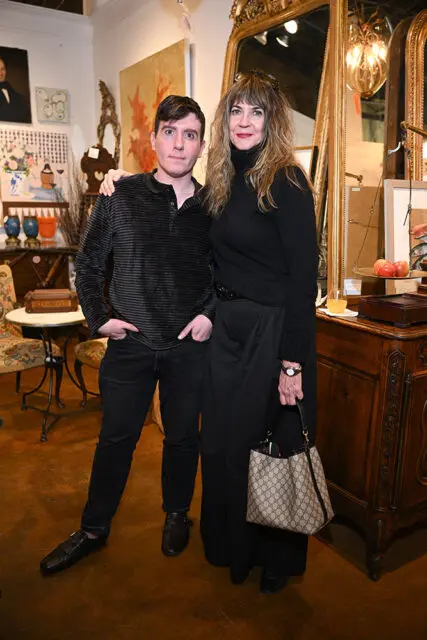 A man and a woman stand together in an antique shop. The man wears a striped polo shirt and dark jeans. The woman wears a black turtleneck and wide-leg pants.