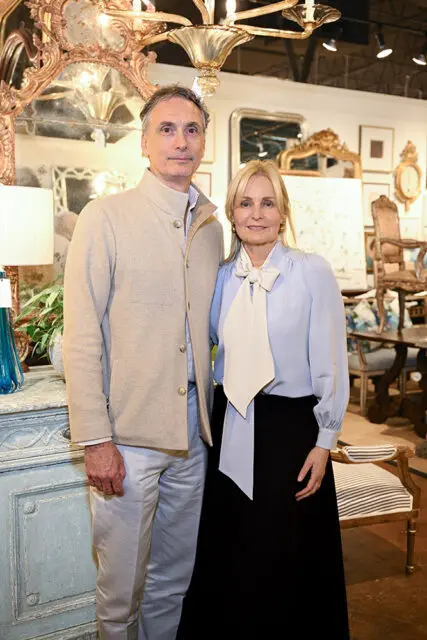 Man in a tan jacket and light pants stands next to a woman in a light blue blouse and black skirt, with antique furniture and mirrors in the background.