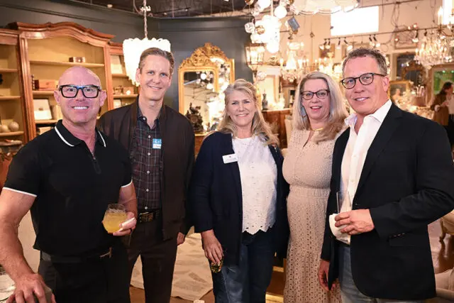 Five people pose for a photo in a showroom filled with chandeliers and antique furniture. The man on the far left wears glasses and a black polo shirt.