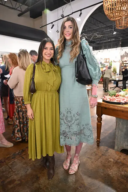 Two women posing for a photo indoors. The woman on the left wears a green dress with a bow tie neck and brown boots. The woman on the right wears a pale blue dress with intricate lace detailing and gold sandals.