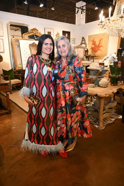 Two women smiling in a room filled with antique furniture and decor. The woman on the left wears a patterned dress with feather trim. The woman on the right wears a floral dress.