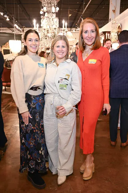 Three women pose for a photo in a store with chandeliers and furniture in the background. The woman on the left wears a tan sweater and floral skirt. The woman in the middle wears a striped jumpsuit and holds a drink. The woman on the right wears an orange ribbed dress.