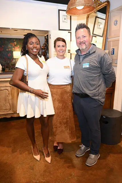 Three people posing for a photo indoors. The woman on the left wears a white dress and holds a drink. The woman in the middle wears a white t-shirt and a brown skirt. The man on the right wears a gray quarter-zip shirt and dark pants.