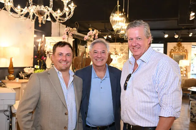 Three men smiling at the camera in a well-lit showroom with ornate chandeliers and decorative mirrors in the background.