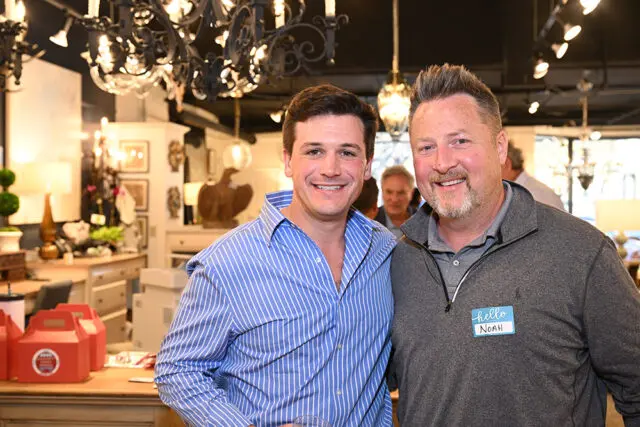 Two men smiling at the camera in a store with chandeliers and decorative items in the background. One man wears a blue and white striped shirt, the other a gray quarter-zip pullover with a "hello NOAH" name tag.