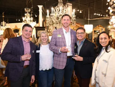 Five people stand together in a showroom filled with chandeliers and furniture. From left to right: a man in a purple plaid blazer, a woman in a white lace top, a man in a lavender plaid blazer with a name tag, a man in a pinstripe blazer, and a woman in a cream textured jacket.