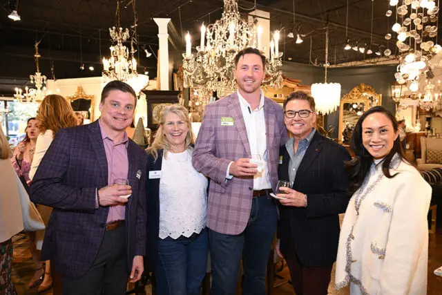 Five people stand together in a showroom filled with chandeliers and furniture. From left to right: a man in a purple plaid blazer, a woman in a white lace top, a man in a lavender plaid blazer with a name tag, a man in a pinstripe blazer, and a woman in a cream textured jacket.