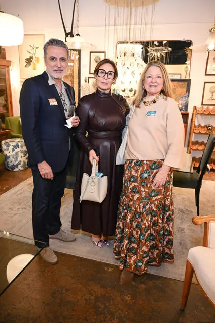 Three people pose for a photo in a room with eclectic decor. The man on the left wears a navy blazer and jeans. The woman in the center wears a dark leather dress and glasses. The woman on the right wears a tan sweater and a patterned skirt.