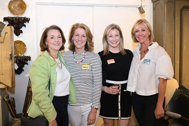 Four women pose for a photo. The woman on the left wears a green jacket and gold necklace. The second woman from the left wears a striped shirt and colorful necklace. The third woman wears a black dress and holds a glass. The woman on the right wears a white shirt with puffy sleeves.