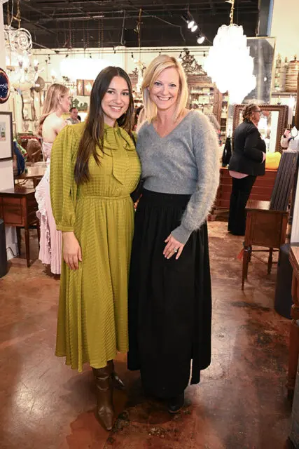 Two women smiling at the camera in an antique store. The woman on the left wears a textured olive green dress and brown boots. The woman on the right wears a gray fuzzy sweater and a long black skirt.