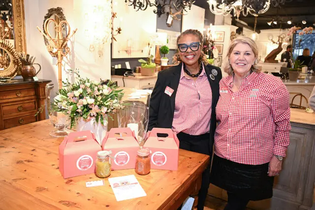Two women in red and white gingham shirts stand behind a table with pink gift boxes and jars.