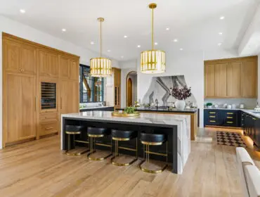Modern kitchen with light wood floors, custom oak cabinetry, a large marble island with black leather bar stools, and brass pendant lights.