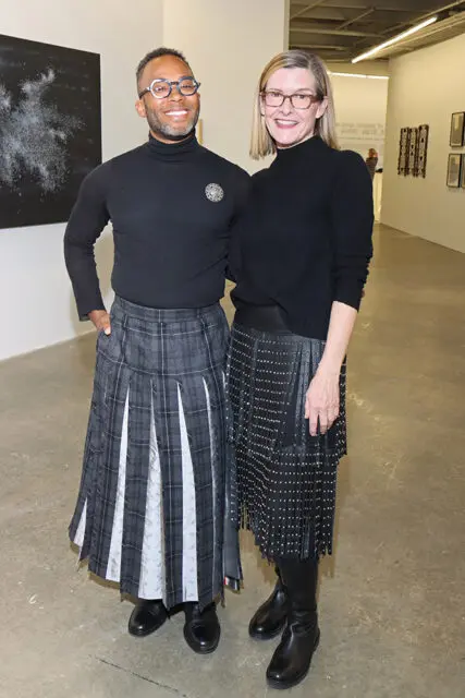 Man in black turtleneck and plaid skirt, and woman in black sweater and studded skirt, stand in an art gallery.