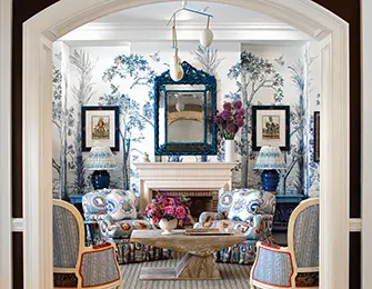 Living room with blue and white Chinoiserie wallpaper, two upholstered armchairs with paisley print, a stone coffee table, and a blue ornate mirror above the fireplace.