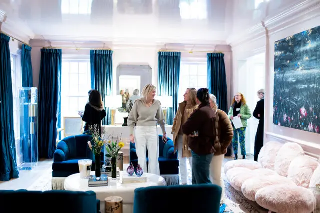 Living room with teal velvet sofas, plush pink sofa, white coffee table, and abstract art on the wall. People are gathered and conversing.